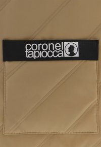 Tejido acolchado beige con costuras diagonales, que presenta una etiqueta negra con texto blanco y una silueta. Detalle de bolsillo en el lado izquierdo.