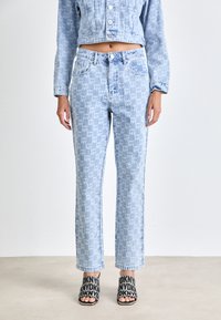 Una chaqueta de mezclilla azul claro y unos vaqueros de cintura alta presentan un patrón de logotipos repetido. Los vaqueros tienen un diseño de pierna recta y se combinan con sandalias negras de estilo deslizante.
