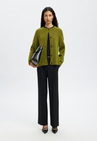 Cárdigan de punto verde con botones negros, llevado sobre una camiseta negra. Combinado con pantalones de pierna ancha negros y tacones puntiagudos negros, sosteniendo un bolso negro.