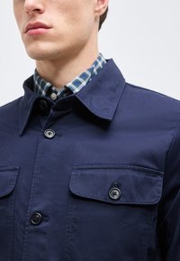 Άνδρας που φοράει μπλε-ναυτικό σακάκι με κουμπιά πάνω από μπλε και πράσινο καρό πουκάμισο με γιακά, εμφανιζόμενος από το πηγούνι έως τον ώμο.