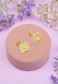 Gyllene blommiga örhängen i form av blommor på en rund, rosafärgad sammetställ. Bakgrunden har små lila och vita blommor mot en pastellfärgad yta.