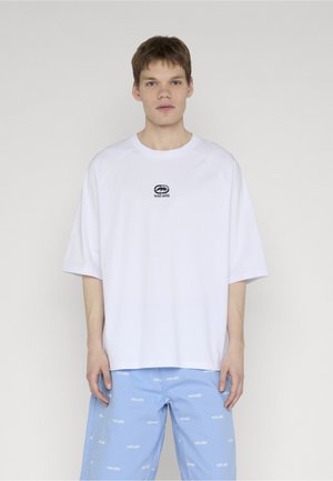 Fiatal férfi túlságosan nagy fehér pólóban, melyen kis fekete logó látható, valamint világoskék rövidnadrágban, amelyen fehér szövegminták vannak.