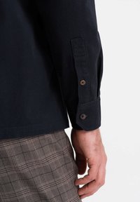 Camisa azul oscura de manga larga, con puños abotonados y una textura suave, acompañada de pantalones de cuadros marrones.