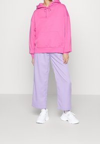 Personne portant un sweat à capuche surdimensionné rose vif, un pantalon large violet clair et des baskets blanches, debout devant un fond uni.