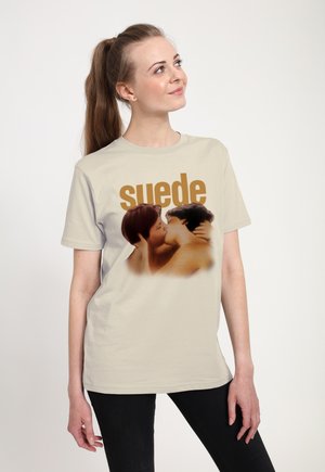 Ung kvinde med lys hud og brunt hår i en hestehale iført en beige t-shirt, der viser to personer, der kysser, og ordet "suede" over dem.