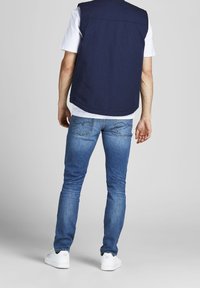 Navy sleeveless vest over a white t-shirt, paired with fitted light blue jeans and white sneakers, viewed from the back.