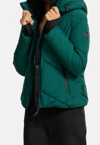 Giacca puffer verde acqua con cappuccio foderato in pelliccia sintetica, chiusura con zip, coulisse regolabile e tasche laterali. Presenta dettagli neri e logo.