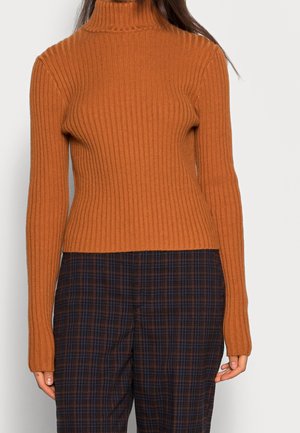 Femme portant un col roulé ajusté en tricot côtelé de couleur orange brûlé et un pantalon taille haute à carreaux foncés, debout devant un fond uni.