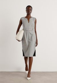 Robe grise à motif avec un décolleté en V, fente sur le côté et taille ceinturée, associée à un sac à main crème et des talons blancs à bout pointu. Texture de tissu lisse.