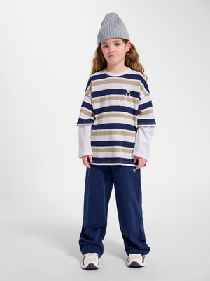 Junges Kind steht und trägt eine graue Mütze, ein gestreiftes beige-navy Shirt über einem weißen Langarmshirt, eine marineblaue Hose und weiße Sneaker vor weißem Hintergrund.