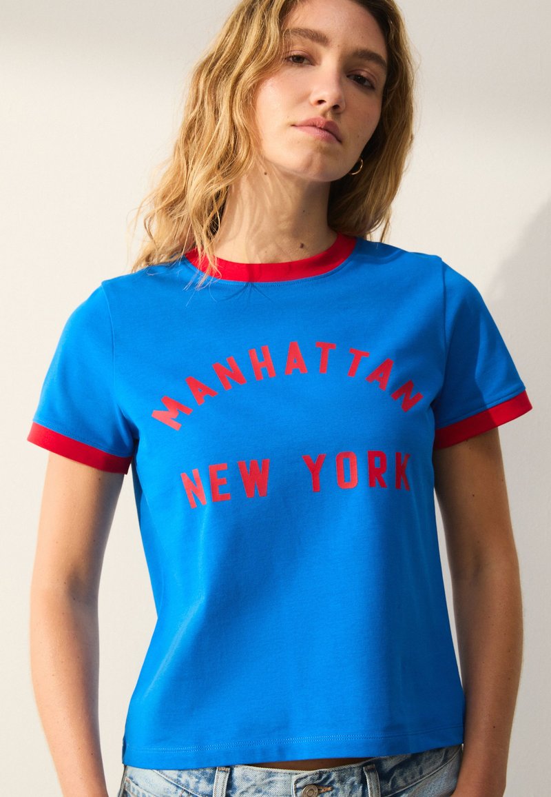 Camiseta de algodón azul con detalles rojos, con "MANHATTAN NEW YORK" en letras rojas llamativas en la parte frontal. Mangas cortas y cuello redondo.