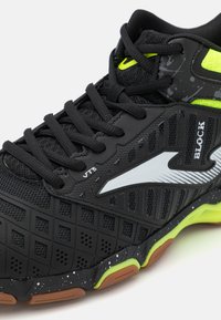Zapatilla deportiva negra con parte superior de malla, detalles de goma texturizada, acentos en verde lima y una suela con un patrón que incluye elementos en naranja y negro moteado.
