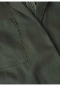 Gros plan sur un tissu vert foncé montrant une poche carrée cousue et une couture avec une fermeture éclair partiellement visible.