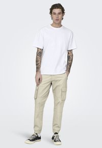 Witte katoenen t-shirt, beige cargo-broek met zijzakken, aansluitende manchetten en zwarte sneakers. Huid met zichtbare tatoeages op de armen.