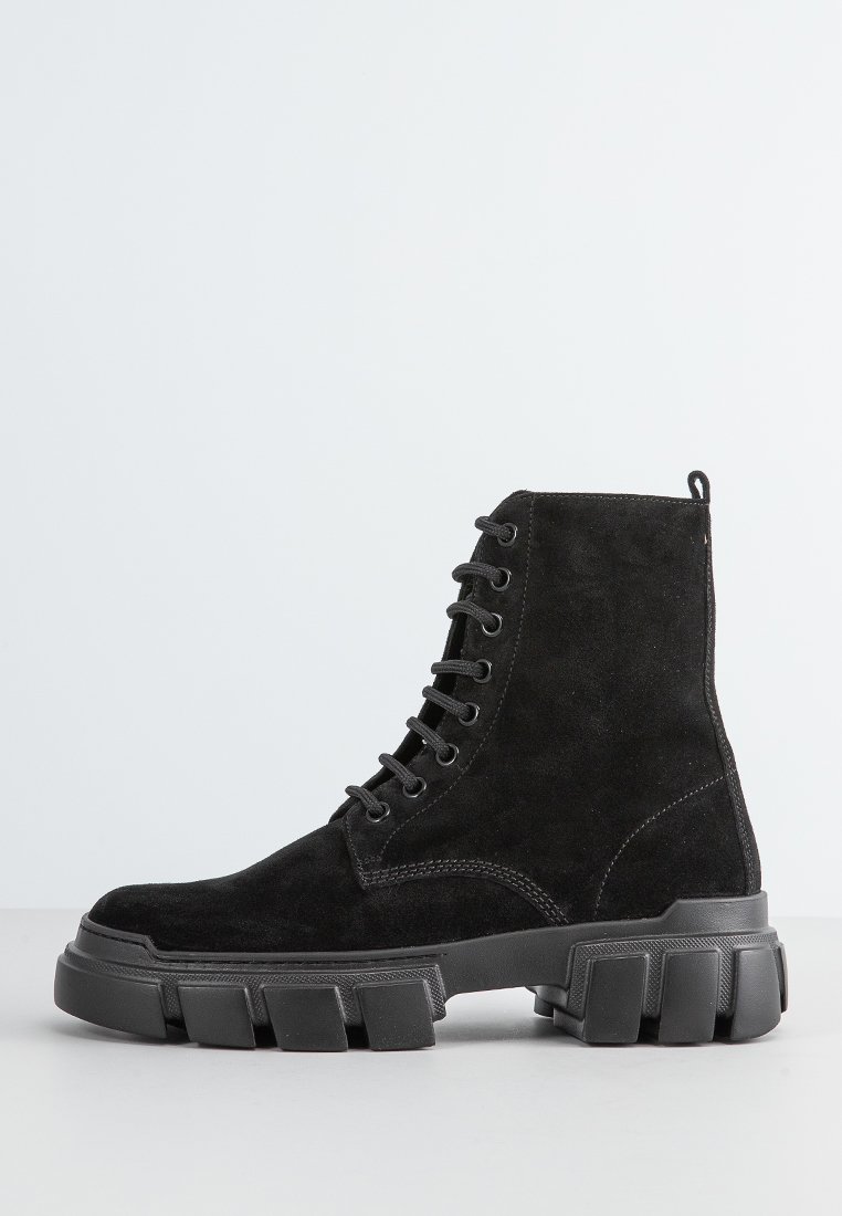 Botas negras de ante hasta el tobillo con una gruesa suela de goma negra, con cordones redondos, puntera redonda y detalles de costura en los laterales.