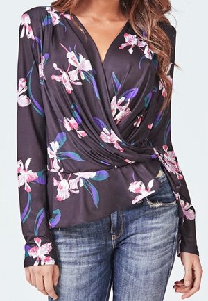 Frau trägt eine schwarze Wickelbluse mit pinken und lila Blumenmustern, kombiniert mit blauen, verwaschenen Jeans.