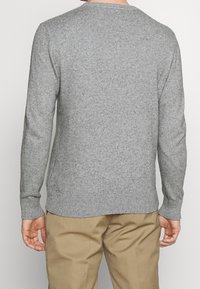 Man in een lichtgrijze trui met lange mouwen en een beige broek, gezien vanaf de achterkant tegen een eenvoudige achtergrond.