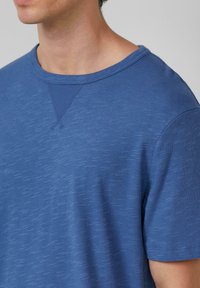 Close-up van een persoon die een blauwe gestructureerde korte mouw shirt met een ronde halslijn en driehoekige stikdetails draagt.