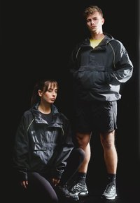 Joven de pie y mujer agachada, ambos vistiendo chaquetas negras y ropa deportiva, contra un fondo negro.