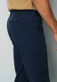 Navy blue trousers made of smooth fabric, featuring a slim fit, side pockets, and a back pocket with a hidden zipper.