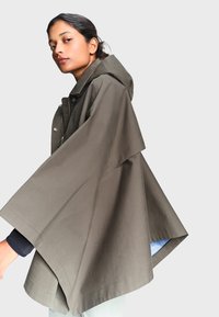 Femme portant une cape manteau à capuche oversize vert olive, posant de profil devant un fond gris clair uni.