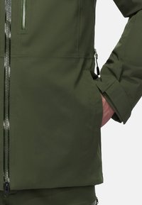 Giacca verde oliva con una finitura liscia e impermeabile. Presenta una tasca con zip, polsini regolabili e una tasca laterale con un cordino visibile.
