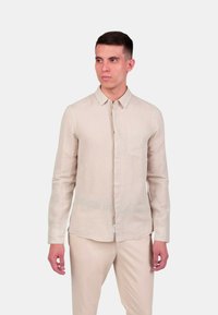 Camicia di lino beige chiaro con design a bottoni, maniche lunghe e taschino sul petto, abbinata a pantaloni di colore chiaro coordinati.