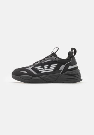Zapatilla deportiva negra y gris con suela gruesa, cordones en la parte delantera, tiradores en la lengüeta y el talón, y logo alado en el lateral.