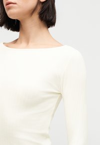 Γυναίκα με κοντά σκουρόχρωμα μαλλιά, που φοράει εφαρμοστό, ριγωτό, μακρυμάνικο λευκό τοπ με φαρδύ λαιμόκοψη, σε απλό φόντο.