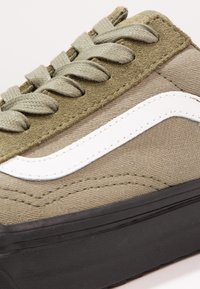 Gros plan sur une basket vert olive et beige avec une bande latérale blanche, des lacets texturés et une semelle noire épaisse.