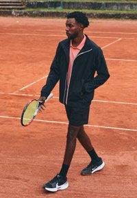 Tenisa spēlētājs melnā jakā ar baltu rāvējslēdzēju, rozā polo krekliņā, melnās šortos un melnās sporta kurpēs, tur krāsainu tenisa racket uz māla laukuma.