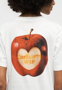 Άτομο που φοράει λευκό μπλουζάκι με μεγάλο κόκκινο γραφικό μήλο στην πλάτη, με μια δαγκωνιά σε σχήμα καρδιάς που αποκαλύπτει θολό κείμενο μέσα.