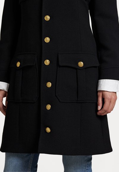 Manteau noir avec deux poches avant, boutons dorés, tissu texturé et design épuré. Les poignets présentent une doublure blanche.