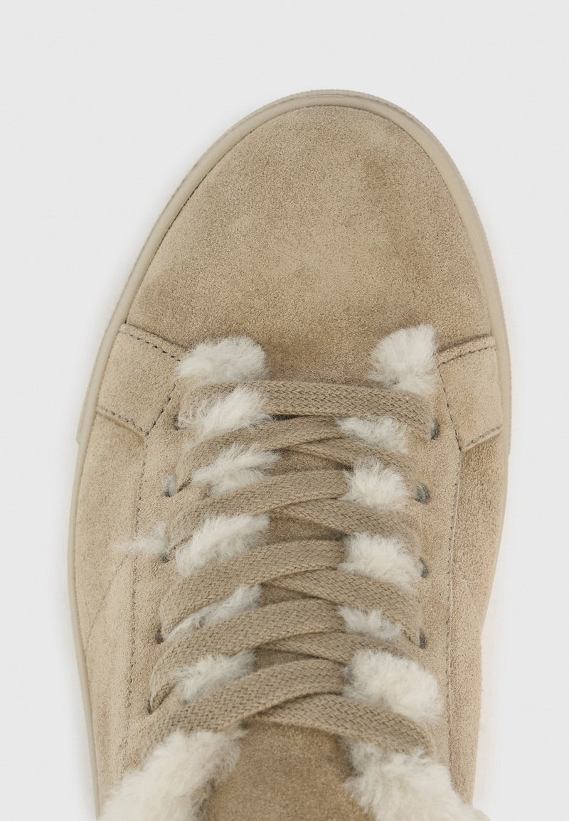 Nahaufnahme eines beigen Wildlederschuhs mit beigen Schnürsenkeln und weißem, flauschigem Futter, das zwischen den Schnürsenkel-Lücken auf einem weißen Hintergrund sichtbar ist.