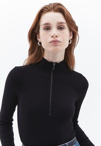 Zwart geribbelde longsleeve top met hoge hals en een ritsdetails aan de voorkant. Glad textuur, aansluitend ontwerp, gecombineerd met een blauwe tailleband.