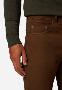 Bruine suède broeken met een textured afwerking, voorzien van voorzakken en gouden hardware. Vergezeld van een donkergroene trui.