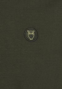 Olivgrön stickad tyg har en cirkulär broderad ugglelogotyp i ljusgrön. Texturen är slät med en lätt stretch.