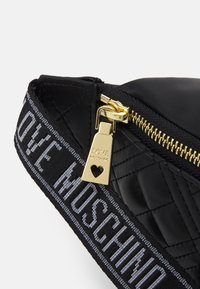 Svart vadderad väska med gulddragkedja och hårdvara. Remmen har upprepande text "LOVE MOSCHINO" i vitt på svart bakgrund.