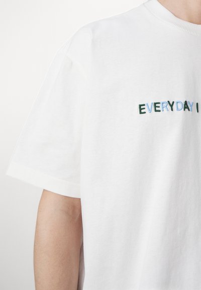 T-shirt en coton blanc à manches courtes avec du texte brodé multicolore "EVERYDAY I" sur le devant. Texture douce.