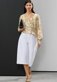 Blouse fluide crème avec un motif floral doré, associée à des culottes blanches amples et des ballerines dorées tressées, complétée par une pochette noire.