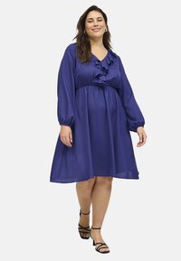 Vestido morado con cuello en V rizado, mangas largas y cintura ajustada. Hecho de una tela ligera, fluye hasta un dobladillo a la altura de la rodilla. Sandalias negras con tiras.