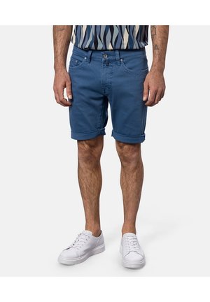 Shorts en denim de coton bleu avec revers roulés, braguette zippée, et design à cinq poches, associés à des baskets blanches et une chemise à motifs.