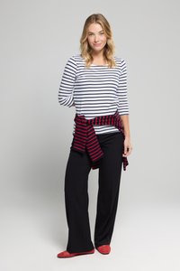 Kvinde med skulderlangt blond hår iført en hvid- og sortstribet top, sorte bukser, en rødstribet sweater knyttet om taljen og røde sko.