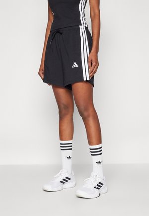 Person trägt schwarze Adidas-Shorts mit weißen Streifen, weiße Adidas-Socken mit schwarzen Streifen und weiße Adidas-Sneakers in einem Studio.