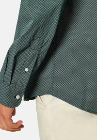 Camisa verde con estampado que presenta una tela texturizada con pequeños diamantes blancos, puños abotonados y una línea de dobladillo curvada.