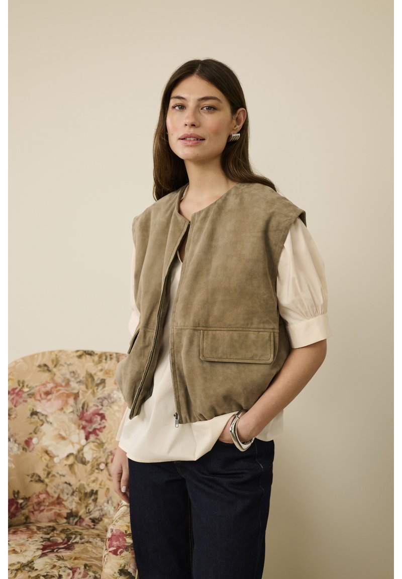 Brun ruskindsvest uden ærmer med rund halsudskæring, lynlås foran og to frontlommer. Båret over en lysefarvet bluse og mørke jeans.