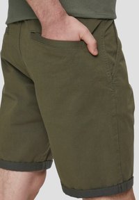 Oliwkowe bawełniane szorty z podwiniętymi brzegami, z bocznymi kieszeniami i gładką teksturą, pokazywane z boku, z ręką w kieszeni.