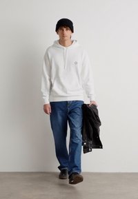 Felpa bianca con tasca frontale e cordino, jeans blu, berretto nero e una giacca di pelle nera appoggiata su un braccio. Scarpe grigie.