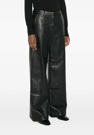 Persoon draagt wijde zwarte leren broek met geweven textuur, gecombineerd met zwarte puntige schoenen en een zwart shirt met lange mouwen, staat stil.