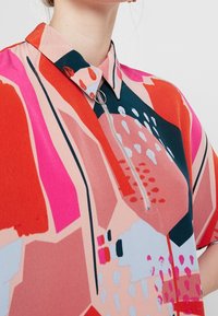 Blouse colorée à manches courtes avec des motifs géométriques dans des nuances de rouge, rose et bleu, dotée d'un col pointu et d'une fermeture éclair à l'avant.